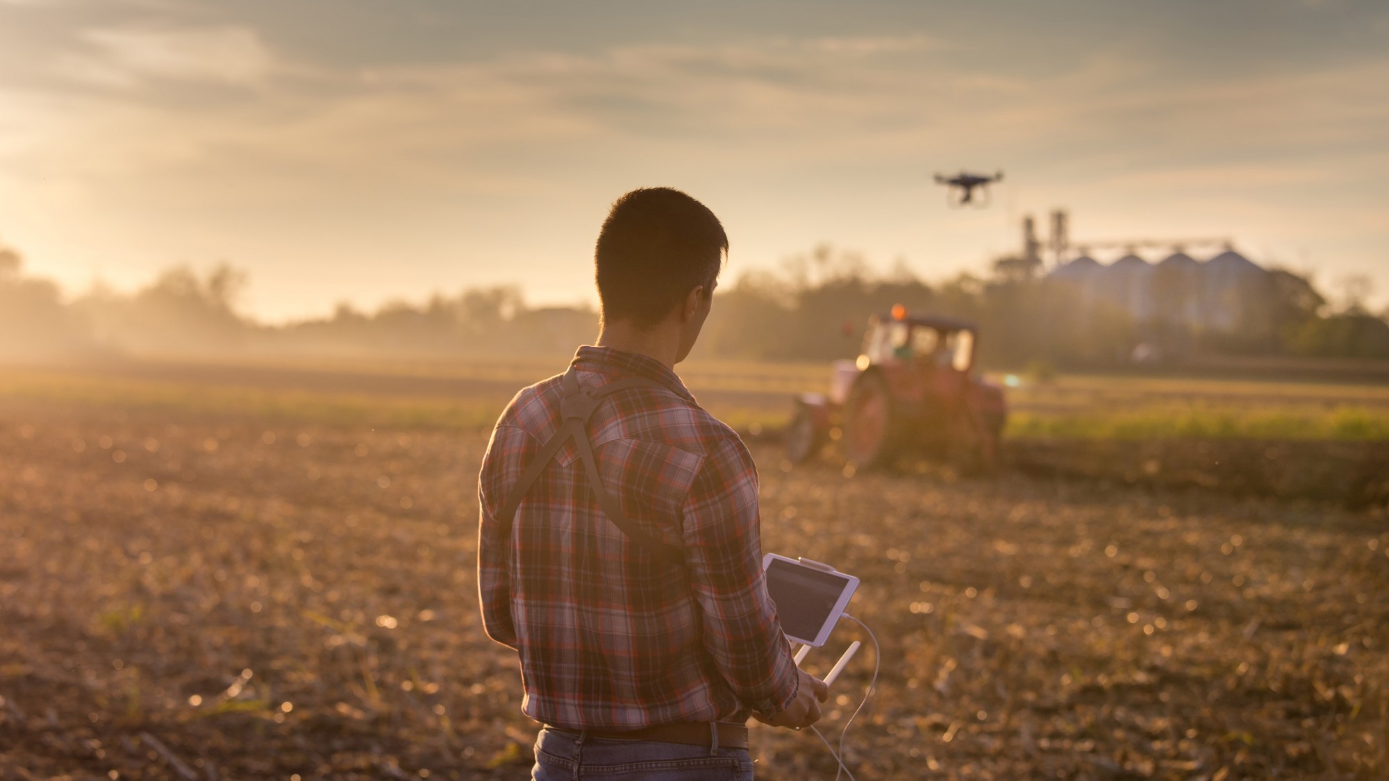 A farmer operates a drone to survey farmland, while a tractor undergoes cultivating operations